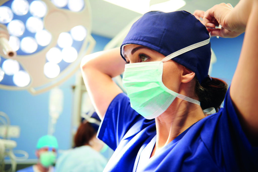 Vue de profil d'une femme accrochant son masque chirurgical dans une salle d'opération de l'Hôpital de Lyon. On aperçoit deux autres personnes appartenant au corps médical de l'hôpital en arrière-plan. 