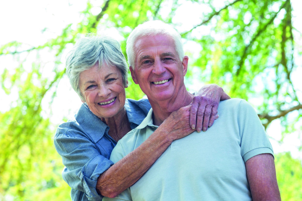 Un couple de senior qui affichent un large sourire pour la photographie. Ils posent à l'extérieur avec de nombreux arbres visibles derrière eux.